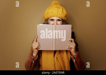 Stilvolle Frau in gelb Baskenmütze und Schal Verstecken hinter Buch auf Bronze Hintergrund. Stockfoto