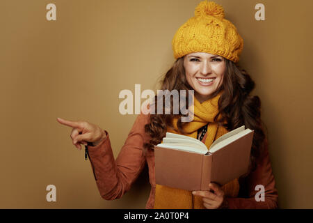 Lächelnd moderne Frau in gelb Baskenmütze und Schal mit Buch an etwas auf beigen Hintergrund zeigt. Stockfoto