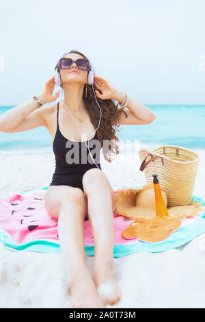 Entspannt trendy 40 Jahre alte Frau mit langen lockigen Haar im eleganten schwarzen Badeanzug an einem weißen Strand Entspannung beim Sitzen auf runde Wassermelonen Handtuch. Stockfoto