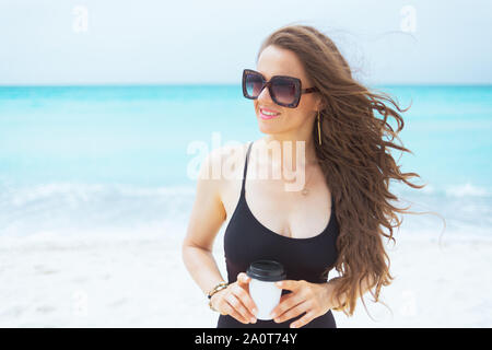 Lächelnd stilvolle mittleren Alter Frau mit langen lockigen Haar in elegantem Schwarz Badeanzug mit Tasse Kaffee an einem weißen Strand. Stockfoto
