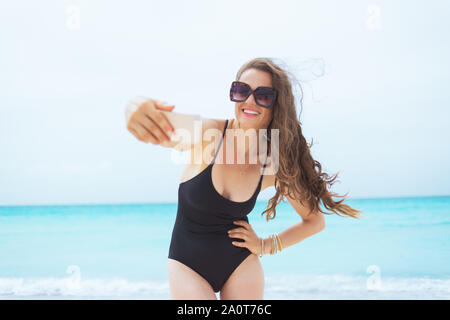 Lächelnden jungen Frau mittleren Alters in Sonnenbrille unter selfie mit Telefon auf einem weißen Strand. Stockfoto