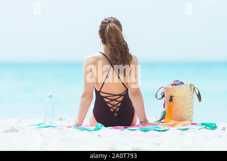 Von hinten trendy mittleren Alter Frau mit langen lockigen Haar in elegantem Schwarz Badeanzug mit Wasserflasche sitzen auf runde Wassermelonen Handtuch auf einen Weißen gesehen Stockfoto