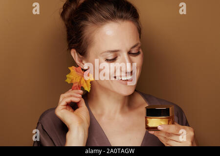 Hallo Herbst. Glücklich moderne 40 Jahre alte Frau in einen Bademantel mit Herbst Blatt Ohrring an Gesichts Creme auf Bronze Hintergrund suchen. Stockfoto