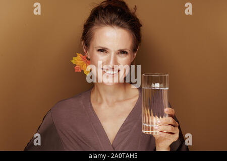 Hallo Herbst. Portrait von Happy Elegant Frau in einen Bademantel mit Herbst Blatt Ohrring mit Glas Wasser auf beige isoliert. Stockfoto