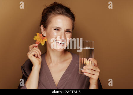 Hallo Herbst. Portrait von Happy Elegant Frau in einen Bademantel mit Herbst Blatt Ohrring holding Glas Wasser auf Bronze Hintergrund isoliert. Stockfoto