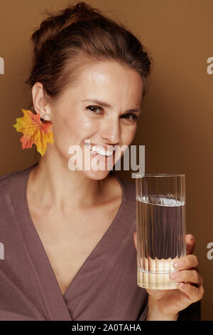 Hallo Herbst. Portrait von lächelnden elegante 40-jährige Frau in einen Bademantel mit Herbst Blatt Ohrring holding Glas Wasser gegen bronze Hintergrund. Stockfoto