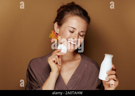 Hallo Herbst. lächelnde junge Frau im Bademantel mit Herbst Blatt mit Gesicht Reinigungsmilch und Wattestäbchen gegen braunen Hintergrund Ohrring. Stockfoto