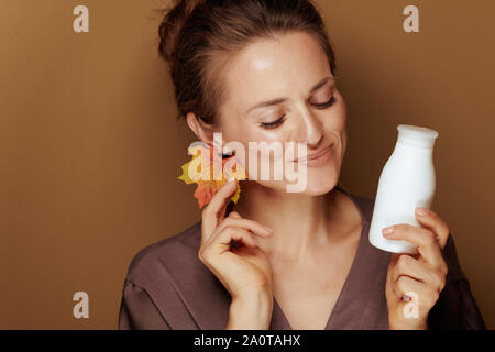 Hallo Herbst. Happy Elegant 40 Jahre alte Frau in einen Bademantel mit Herbst Blatt Ohrring und Gesicht Reinigungsmilch auf Bronze Hintergrund. Stockfoto