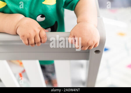 In der Nähe von Babys Hände während des Aufstehens und halten auf dem Geländer der Krippe. Das Tragen eines niedlichen grünen T-Shirt in einem hellen minimalistischen modernen nurs Stockfoto