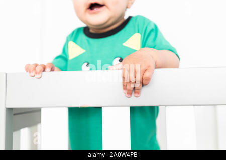 In der Nähe von Babys Hände während des Aufstehens und halten auf dem Geländer der Krippe. Das Tragen eines niedlichen grünen T-Shirt in einem hellen minimalistischen modernen nurs Stockfoto