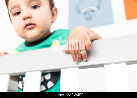 In der Nähe von Babys Hände während des Aufstehens und halten auf dem Geländer der Krippe. Das Tragen eines niedlichen grünen T-Shirt in einem hellen minimalistischen modernen nurs Stockfoto