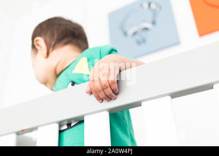 In der Nähe von Babys Hände während des Aufstehens und halten auf dem Geländer der Krippe. Das Tragen eines niedlichen grünen T-Shirt in einem hellen minimalistischen modernen nurs Stockfoto