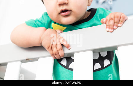 In der Nähe von Babys Hände während des Aufstehens und halten auf dem Geländer der Krippe. Das Tragen eines niedlichen grünen T-Shirt in einem hellen minimalistischen modernen nurs Stockfoto