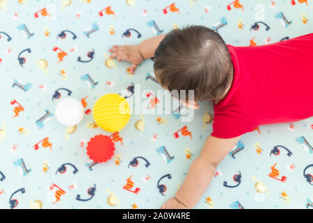 Baby spielt mit bunten Spielzeug Gummi Kugeln in der Krippe in einem hellen Raum. Kind trägt einen roten Strampler, Verlegung auf spielerische Dinosaurier Krippe Blatt. Stockfoto