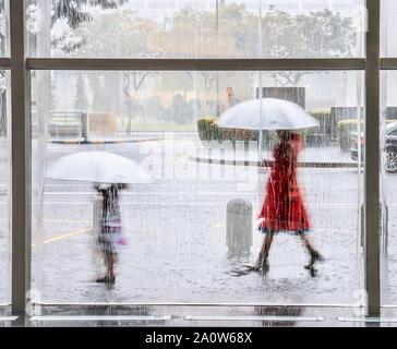 Ein tropischer Monsunregen in Singapur gesehen durch ein Fenster mit Wasser streifig ist, als Mutter und Tochter zu Fuß außerhalb mit passenden weißen Sonnenschirmen. Stockfoto