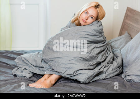 Zufrieden zufrieden Frau sitzt mit warmen weichen grauen Decke in ein bequemes Bett. Schönes verschlafenes weibliche Ausgabe Zeit im Schlafzimmer. Erholen, Entspannen, Wee Stockfoto