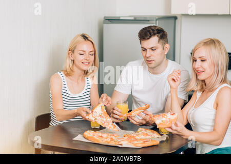 Drei kaukasischen gerne Studenten essen Pizza, trinken Orangensaft in der Küche. Freunde in Partei zu Hause, reden, lachen und Spaß haben. Freunde Stockfoto