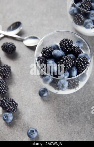 Selektiver Fokus der leckeren Joghurt mit Chia Samen, Heidelbeeren und Brombeeren in Gläsern in der Nähe von Teelöffel und zerstreute Beeren auf Marmor Oberfläche Stockfoto