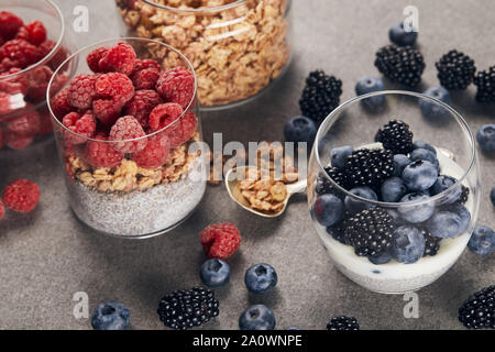 Selektiver Fokus der leckeren Joghurt mit Chia Samen, Beeren und Haferflocken in Gläsern in der Nähe von Teelöffel auf Marmor Oberfläche Stockfoto