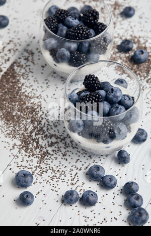 Selektiver Fokus der leckeren Joghurt mit Chia Samen, Blaubeeren, Brombeeren in der Nähe von verstreuten Samen und Beeren auf weißem Holz- Stockfoto
