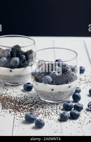 Selektiver Fokus der leckeren Joghurt mit Chia Samen, Blaubeeren, Brombeeren in der Nähe von verstreuten Samen und Beeren auf weißem Holz isoliert auf Schwarz Stockfoto