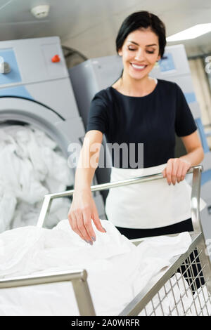 Selektiver Fokus der glücklichen Hausmädchen in der Nähe von Korb mit weißen Bettlaken in Wäscherei Stockfoto