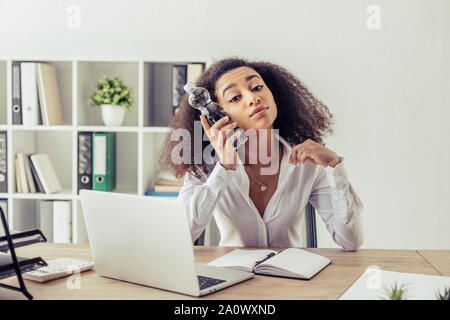 Ziemlich afrikanische amerikanische Geschäftsfrau auf Kamera, während am Arbeitsplatz sitzen und halten Plastikflasche in der Nähe von Kopf Stockfoto