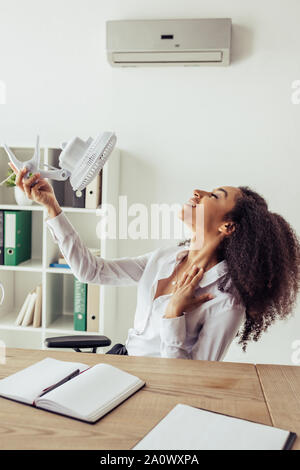 Ziemlich afrikanische amerikanische Geschäftsfrau am Arbeitsplatz sitzen und Betrieb elektrischer Ventilator beim Sitzen am Arbeitsplatz im Büro Stockfoto