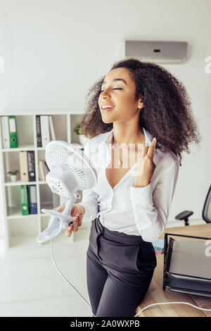 Ziemlich afrikanische amerikanische Geschäftsfrau, die in der Nähe von Arbeitsplatz und Betrieb elektrischer Ventilator Stockfoto