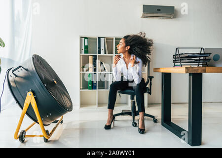 Ziemlich afrikanische amerikanische Geschäftsfrau, sitzen auf einem Bürostuhl vor Blasen elektrischer Ventilator Stockfoto