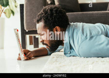 Afrikanische amerikanische Mann auf der Suche nach Thermometer beim liegen auf dem Boden und das Leiden von der Sommerhitze schockiert Stockfoto