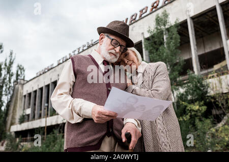 PRIPYAT, UKRAINE - 15. AUGUST 2019: Low Angle View der älteren Pärchen auf der Suche nach Foto in der Nähe von Gebäude in Tschernobyl. Stockfoto