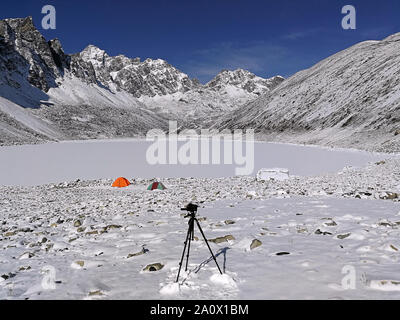 Kamera auf einem Stativ auf dem Hintergrund der Zeltlager auf der Bank eines zugefrorenen See in die Berge des Himalaja; Tourismus und Reisen Konzept Stockfoto