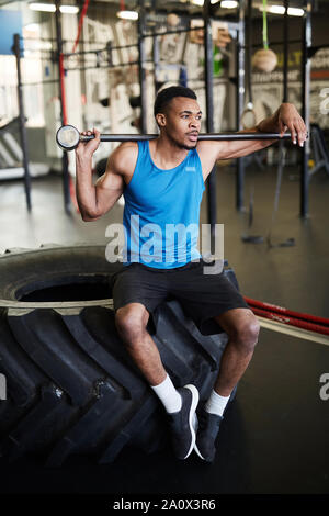 In voller Länge Porträt der muskulösen Afrikaner - der Mann sitzt auf Reifen und halten Hammer während der Pause von Cross Training im modernen Fitnessraum Stockfoto