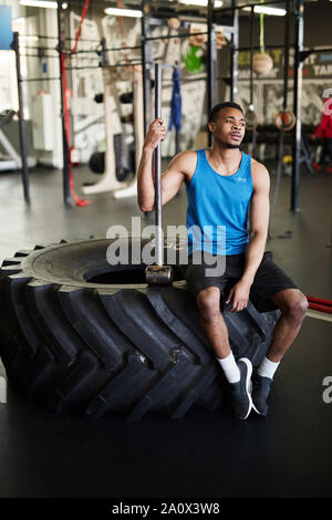 In voller Länge Porträt der muskulöse Afrikanische Mann sitzt auf Reifen und halten Hammer während der Pause von Cross Training im modernen Fitnessraum Stockfoto