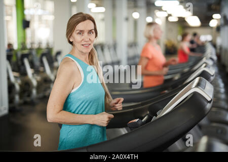 Taille bis Portrait von reife Frau an der Kamera schaut während der Ausführung auf Laufband während cardio Workout im modernen Fitnessraum, Kopie Raum Stockfoto
