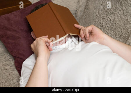 Ein Mann in einem weißen T-Shirt ist das Schlafen auf einem Sofa mit einem Buch auf seinem Gesicht. Stockfoto
