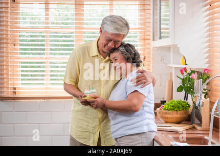 Asiatische älterer Mann überraschen ältere Frau mit Geburtstag Kuchen in der Küche zu Hause. umarmen und genießen die Zeit togethe, Altern zu Hause Konzept Stockfoto