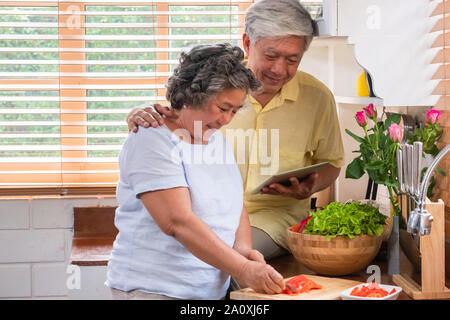 Asiatische senior Frau schneiden Tomaten auf Holz Schneidebrett und mit Tablet-PC zu suchen Menü Rezept. Senior mit Technologie lfiestyle. Altern Stockfoto