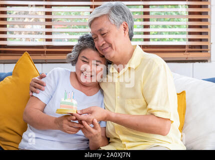 Asiatische älterer Mann überraschen ältere Frau mit Geburtstag Kuchen im Wohnzimmer zu Hause. Altern zu Hause Konzept. Stockfoto