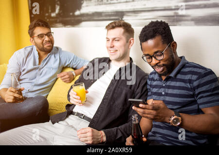 Junge afrikanische amerikanische bärtiger Mann mit Handy, während Sie Filme oder Sport Spiele mit Freunden. Der Gruppe multikulturellen Studenten Spaß a Stockfoto