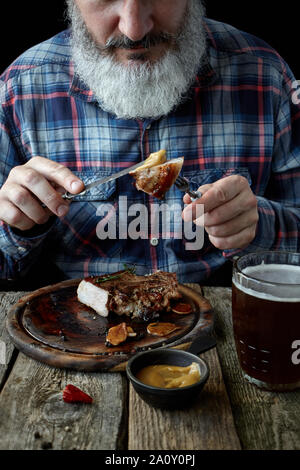 Nahaufnahme der brutalen Grauhaarige erwachsenen Mann mit einem Bart Senf Steak isst und trinkt Bier, das Konzept eines Urlaubs, Festival, Oktoberfest oder St. Patrick's Da Stockfoto