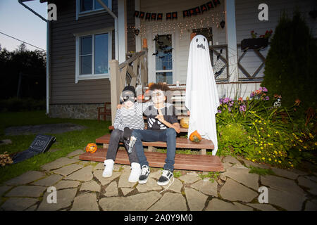 In voller Länge Porträt von zwei jungen tragen Halloween Kostüme posiert im Freien auf der Veranda sitzen zusammen in dunklen, geschossen mit Flash, kopieren Raum Stockfoto