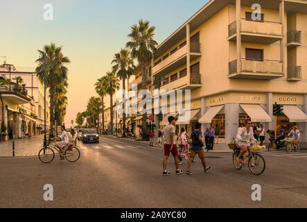 Blick auf das Zentrum von Forte dei Marmi mit Fußgänger und Radfahrer beim Überqueren einer von Palmen gesäumten Straße bei Sonnenuntergang, Lucca, Toskana, Versilia, Italien Stockfoto