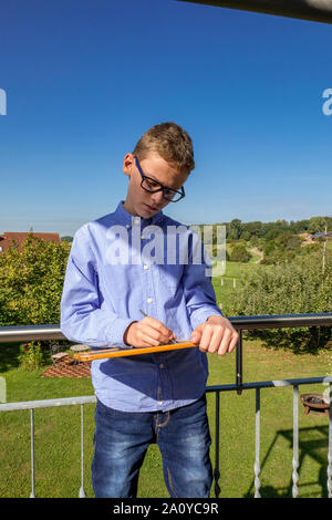 Junge mit Brille steht auf dem Balkon und Schreibt etwas auf einer kleinen Tafel Stockfoto