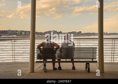 Senior Paar von hinten sitzen auf einer Bank der Pontile Bellavista Vittoria mit dem Stadtbild bei Sonnenuntergang im Sommer, Lido di Camaiore, Toskana, Italien Stockfoto