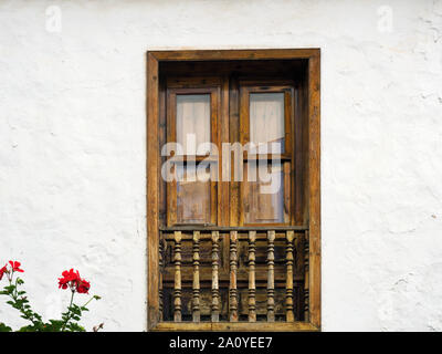 Typische Holz- Fenster auf den Kanarischen Inseln mit gedrehten Holzpfähle dekoriert auf einem weißen Hauswand in Nahaufnahme. In der unteren linken, eine Pflanze mit roten Stockfoto