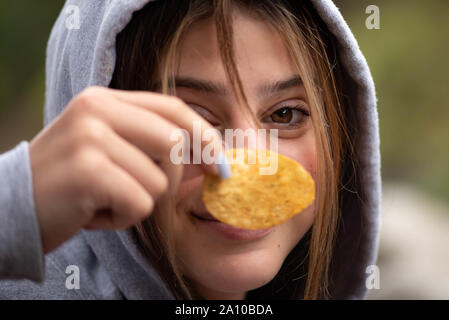 Nahaufnahme von einem jungen Mädchen lächelnd, Holding a Potato Chip vor ihrem Gesicht, trug einen Kapuzenpulli.. Stockfoto