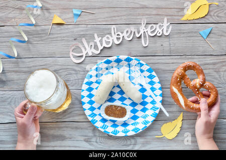 Oktoberfest, traditionellen Festival Essen: Weißwurst, Hände, die Brezel und Bier. Flach auf rustikalen Holztisch mit blau und weiß Dekoration Stockfoto