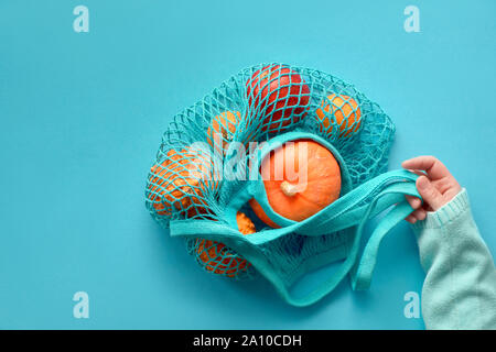 Herbst flach mit weiblichen Hand türkis String Tasche mit orange Kürbisse. nach oben Blick auf Papier Hintergrund in blau mint Farbe. Stockfoto